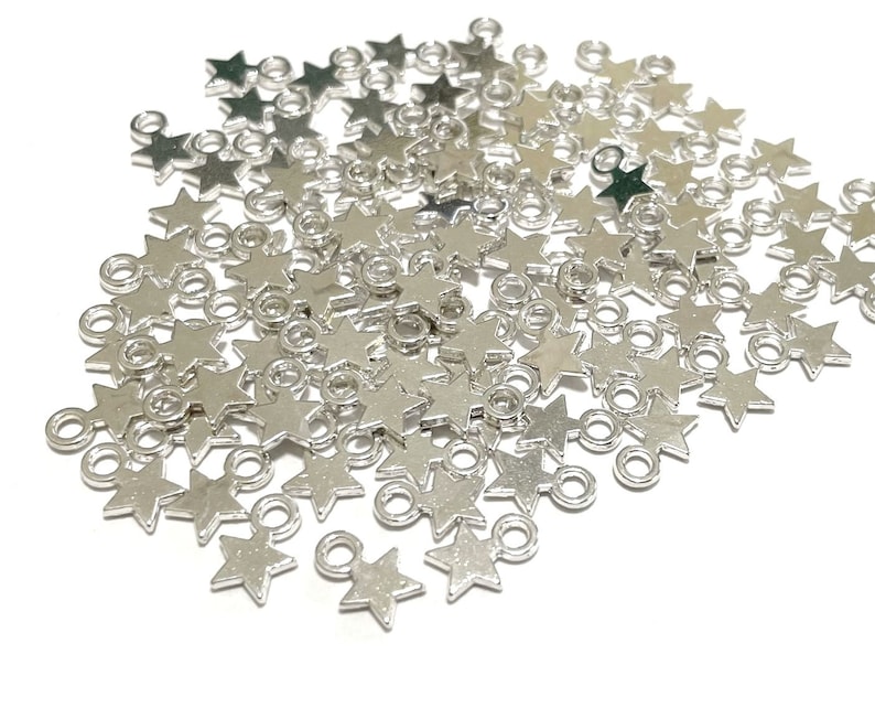 May include: A large pile of silver star-shaped charms with a jump ring attached to each star. The charms are scattered on a white background.