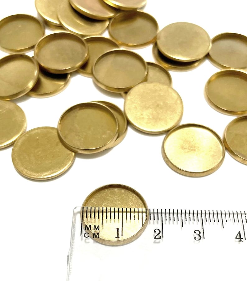 Puede incluir: Un mont&oacute;n de 20 discos redondos de metal de color dorado, cada uno con un borde elevado. Una regla se coloca junto a uno de los discos, mostrando que el disco tiene un di&aacute;metro de 2,5 cm.