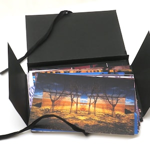 May include: A black photo album with a ribbon closure. The album is open and filled with colorful photographs. The top photo shows a landscape with trees and a sunset.