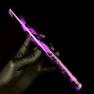May include: A cylindrical object with a swirling purple and black pattern. The object is held against a black background. The colors create a marbled effect, with the purple dominating the design. The object appears to be made of a solid material.
