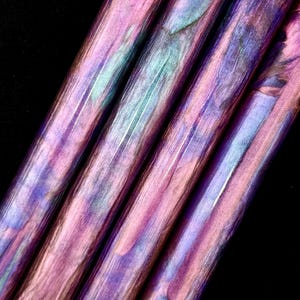 Puede incluir: Primer plano de varios objetos cilíndricos con apariencia marmolada. La paleta de colores incluye tonos de rosa, morado y verde azulado, creando un efecto visual llamativo. Los objetos están dispuestos en diagonal sobre un fondo negro.
