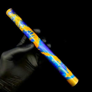 May include: A cylindrical object with a vibrant, swirling pattern of blue, gold, and yellow hues. The object is held against a black background. The colors blend together, creating a marbled effect. The ends have a gold finish.