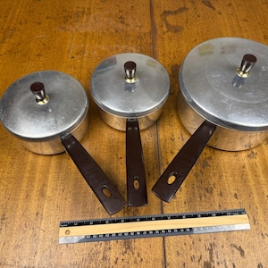 May include: A set of three silver-colored aluminum saucepans with dark brown handles and matching lids, each featuring a central brown knob. A wooden ruler is positioned below the pans, showing measurements in inches and centimeters for scale.