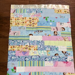 May include: A patchwork baby blanket with a satin border. The blanket features various patterned fabrics in pastel colors, including floral, animal, and geometric designs. The blanket is rectangular and appears to be handmade.