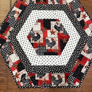 May include: Hexagon-shaped quilted table runner with a rooster theme. The runner features a patchwork design with red, black, white, and tan fabrics. The center has white fabric with black polka dots. The outer edges have a black and white triangle pattern.