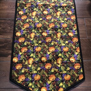 May include: A quilted table runner with a harvest theme. The runner features a repeating pattern of orange pumpkins, purple grapes, and green leaves on a dark background. The edges are trimmed with a black and white polka dot border.