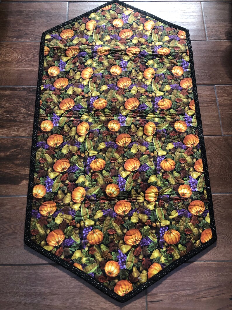 May include: A quilted table runner with a harvest theme. The runner features a repeating pattern of orange pumpkins, purple grapes, and green leaves on a dark background. The edges are trimmed with a black and white polka dot border.
