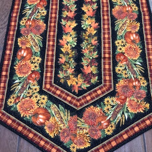 May include: A quilted table runner with a black background, featuring autumn-themed designs. The runner showcases pumpkins, sunflowers, and fall leaves in shades of orange, yellow, and red. Plaid borders frame the central design.