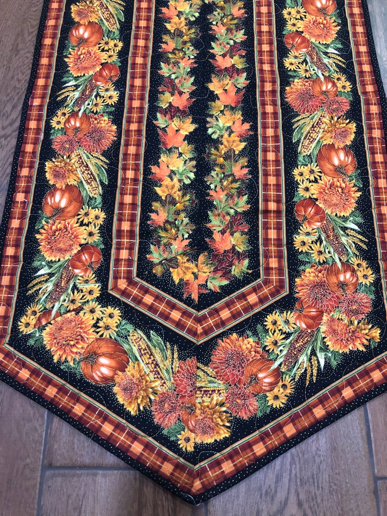 May include: A quilted table runner with a black background, featuring autumn-themed designs. The runner showcases pumpkins, sunflowers, and fall leaves in shades of orange, yellow, and red. Plaid borders frame the central design.