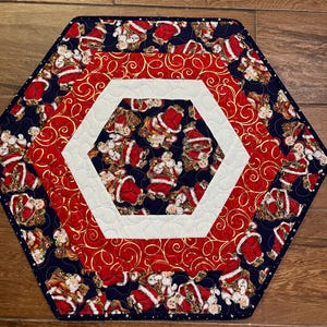 May include: Hexagon-shaped quilted table runner with a Christmas theme. The runner features a navy blue border with Santa Claus figures, a red center with gold swirls, and a white hexagon in the center. The runner is approximately 20 inches in diameter.