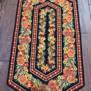 May include: A quilted table runner with a black background and a fall harvest theme. The runner features pumpkins, sunflowers, and autumn leaves. The border has a plaid pattern in shades of orange and brown.