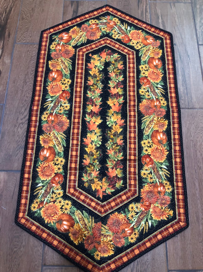 May include: A quilted table runner with a black background and a fall harvest theme. The runner features pumpkins, sunflowers, and autumn leaves. The border has a plaid pattern in shades of orange and brown.