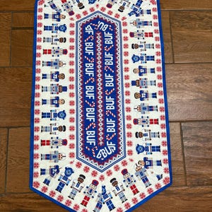 May include: A decorative table runner featuring a repeating pattern of football players in blue, red, and white uniforms. The center panel has the word "BUF" in a repeating pattern. The runner has a blue border and red snowflake accents.
