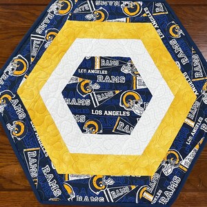 May include: Hexagon-shaped quilted fabric coaster featuring the Los Angeles Rams football team logo and colors. The design includes navy blue, yellow, and white fabric with the team name and logo.