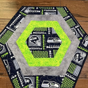 May include: Hexagon-shaped quilted table mat featuring the Seattle Seahawks logo and colors. The mat has a navy blue border with the team name and logo, a gray middle section, and a bright green center with the Seahawks logo. The mat is approximately 15 inches across.
