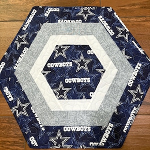 May include: Hexagon-shaped quilted table runner featuring a navy blue fabric with white stars and the word "COWBOYS." The runner has a white and gray border. The design is a tribute to the Dallas Cowboys football team.
