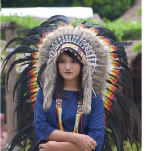 May include: A woman wearing a large black and red feathered headdress with a beaded band. She is wearing a blue shirt and a beaded necklace.