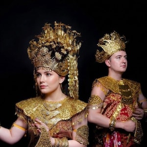Könnte beinhalten: Zwei Personen in traditioneller indonesischer Kleidung, mit aufwendigen goldenen Kopfbedeckungen und passenden goldenen Schulterstücken. Die Kostüme umfassen rot-goldene Stoffe mit Mustern und aufwendigen Goldschmuck. Der Hintergrund ist schwarz.