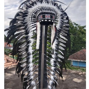 Peut inclure: Coiffe cérémonielle noire et blanche. La coiffe est ornée de nombreuses plumes, et la zone centrale de la couronne présente des perles détaillées rouges, noires et blanches. Les plumes sont disposées symétriquement.