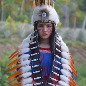 May include: A child wearing a traditional Native American headdress with white feathers and orange accents. The headdress has a black leather band with white beads and a beaded fringe. The child is wearing a blue and red shirt with a geometric pattern.