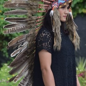Peut inclure: Une femme portant un haut en dentelle noire avec une coiffe en plumes brunes et blanches. La coiffe a un bandeau rouge, blanc et bleu avec un motif d'étoile.