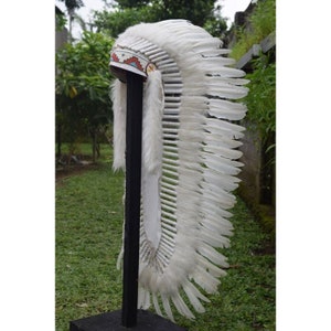 May include: A white feather headdress with a beaded band and white fur trim. The headdress is mounted on a black stand.