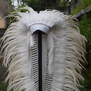 Könnte beinhalten: Ein weißer Federkopfschmuck mit einem weißen Perlenband und einer weißen Filzkrone. Der Kopfschmuck wird auf einem schwarzen Ständer ausgestellt.
