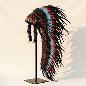 Op de afbeelding: Een hoge, uitgebreide hoofdtooi met een lange sleep van zwarte veren met felblauwe en rode accenten. De voorband is versierd met een geometrisch patroon in oranje, wit en zwart, afgewerkt met ronde zijmedaillons en zwart bont.