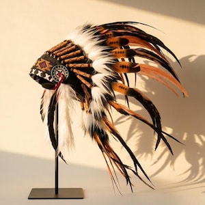 Op de afbeelding: Een decoratieve hoofdtooi met lange, gelaagde veren in bruine, zwarte en witte tinten. De band is versierd met ingewikkeld geometrisch kraalwerk in oranje, zwart en wit, geaccentueerd door een rond embleem met een centraal sterontwerp.