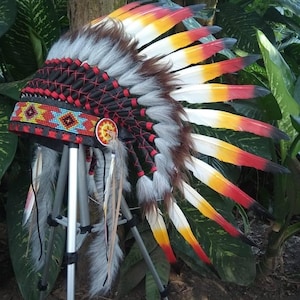 May include: A Native American headdress with a black, red, and gray beaded headband. The headdress features a central medallion and is adorned with white, yellow, and red feathers. The headdress is displayed on a tripod.