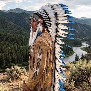 Op de afbeelding: Een persoon draagt een traditionele Native American hoofdtooi met witte, zwarte en blauwe veren. De persoon draagt ook een bruin suède jasje met franjes en kralen, en houdt een houten staf vast. De achtergrond toont een rivier en bergen.