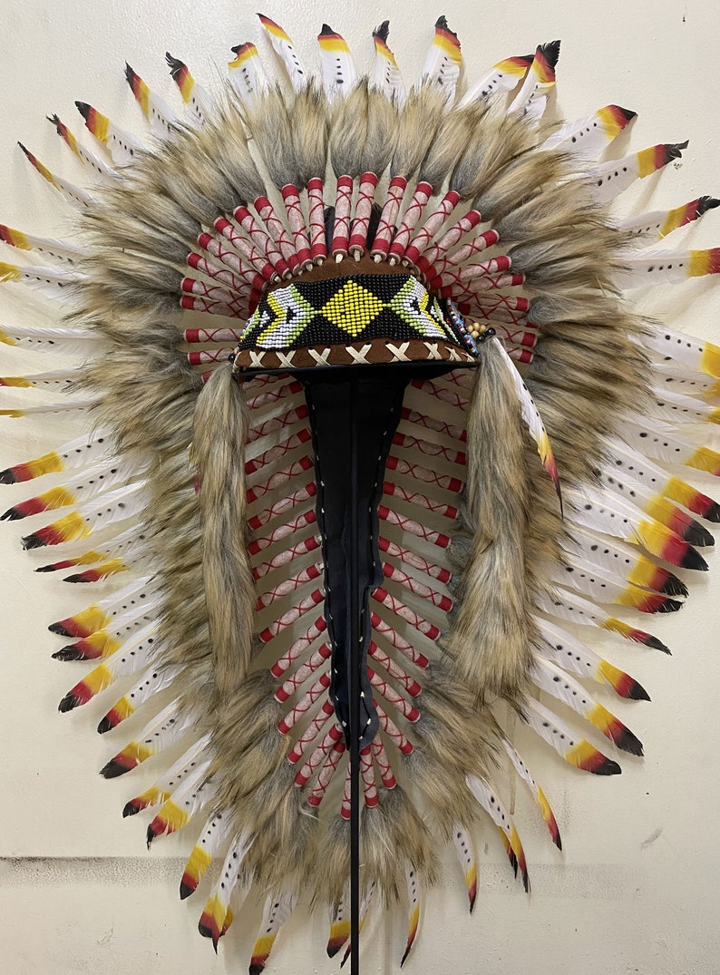 May include: A traditional Native American headdress with white and yellow feathers, red and white wrappings, and a brown beaded band with yellow and white geometric designs.