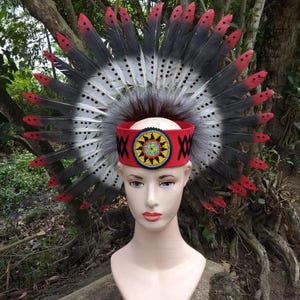 Pode incluir: Um cocar cerimonial com uma faixa de feltro vermelho adornada com um desenho de contas e padrões 'X' pretos. O cocar é adornado com penas cinzentas, pretas e vermelhas. As penas irradiam da faixa.