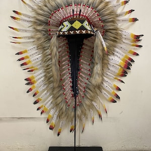 May include: A Native American headdress with white, brown, and red feathers, a beaded band, and a brown base. The headdress is displayed on a black stand.
