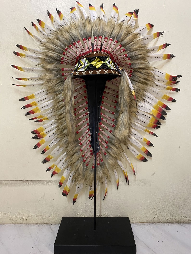 May include: A Native American headdress with white, brown, and red feathers, a beaded band, and a brown base. The headdress is displayed on a black stand.