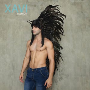 May include: A person wearing a black feather headdress and blue jeans. The headdress is tall and has many feathers. The person is standing in front of a grey wall.