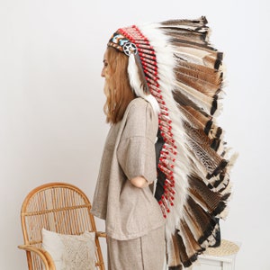 May include: A woman wearing a brown linen top and pants with a large, colorful Native American headdress made of feathers.