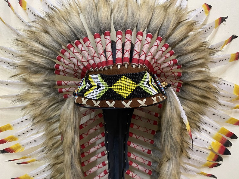 May include: A brown and black beaded headdress with white and yellow feathers. The headdress has a brown leather band with a beaded design and a red and white fringe.