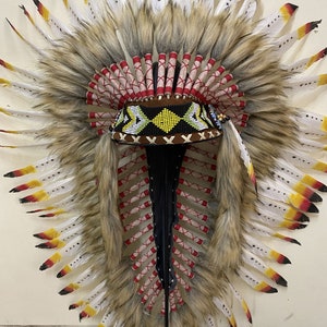 May include: A Native American headdress with white and yellow feathers, red and white trim, and a beaded band. The headdress is mounted on a black stand.