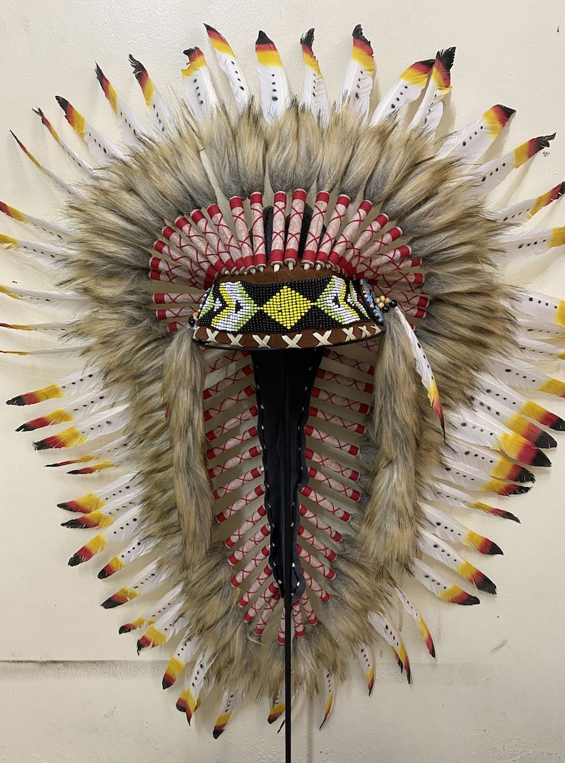 May include: A Native American headdress with white and yellow feathers, red and white trim, and a beaded band. The headdress is mounted on a black stand.