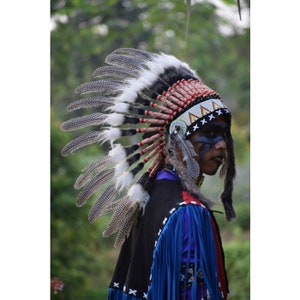 Pode incluir: Uma pessoa usando um cocar tradicional nativo americano com penas, contas e uma faixa vermelha e branca. O cocar é feito de penas brancas e tem uma guarnição de pele preta e branca. A pessoa está usando uma camisa azul e preta com franjas.