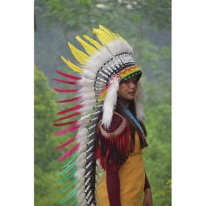 May include: A person wearing a colorful Native American headdress with white, yellow, red, and green feathers. The headdress has a beaded band and a red star on the front. The person is wearing a yellow dress and a red shirt.