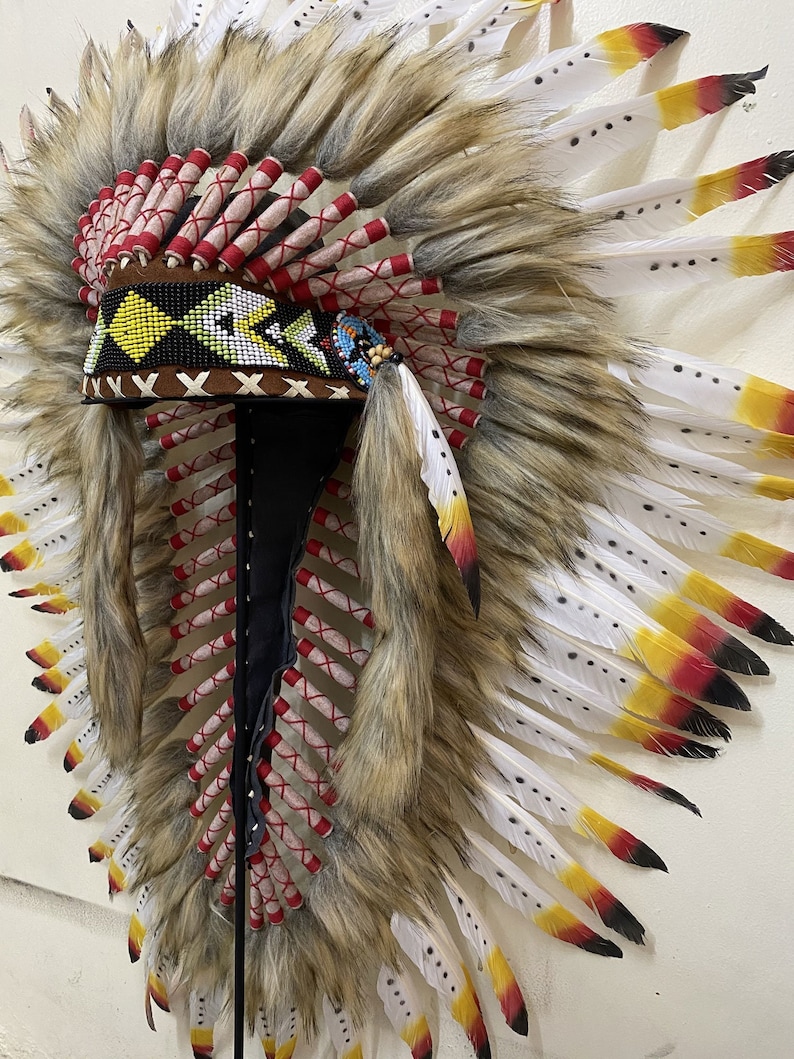 May include: A Native American headdress with white feathers, red and yellow accents, and a beaded band. The headdress is made of feathers, fur, and beads.