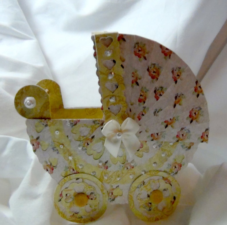 May include: A yellow and white baby carriage with a floral pattern and a white satin bow. The carriage is made of cardboard and has a scalloped edge.
