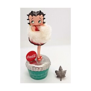 May include: A Betty Boop figurine wearing a red dress and a white fur wrap, standing on a green and silver Coca-Cola bottle cap base. The base has the words "Delicious and Refreshing". A small silver leaf is next to the figurine.