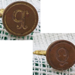 以下が含まれることがあります： 白い緑の布地の背景に、レリーフの文字「G」と「Q」が刻まれたアンティークな真鍮製のレターシールが2つあります。