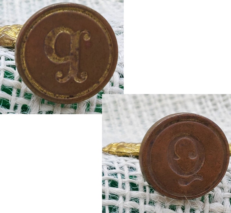 以下が含まれることがあります： 白い緑の布地の背景に、レリーフの文字「G」と「Q」が刻まれたアンティークな真鍮製のレターシールが2つあります。