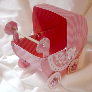 May include: A pink and white baby carriage made of paper with a white lace design and pink and white stripes. The carriage has a white ribbon and a pink bow on the back.