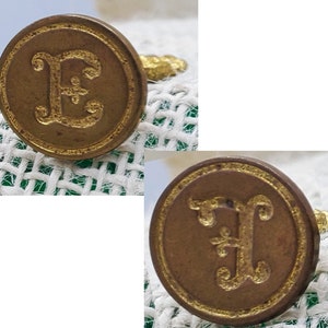 以下が含まれることがあります： 装飾的なフォントで金色の文字「E」が刻まれた、アンティークな真鍮製のワックスシールが2つ。文字「E」の中央には小さな花柄のデザインが施されています。