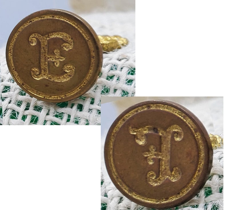 以下が含まれることがあります： 装飾的なフォントで金色の文字「E」が刻まれた、アンティークな真鍮製のワックスシールが2つ。文字「E」の中央には小さな花柄のデザインが施されています。